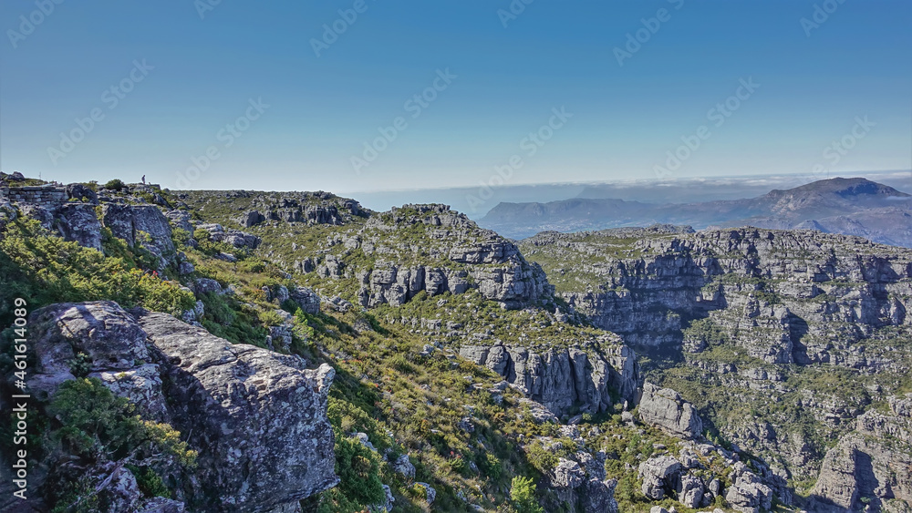 A tiny silhouette of a man is visible on the flat top of Table Mountain. There is scant vegetation on the cracked gray stones. Blue sky. Cape Town. South Africa