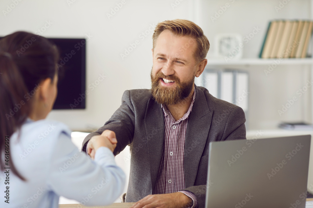 Happy handsome bearded man in suit shaking hands with young woman at business meeting. Smiling client exchanging handshake with bank manager. Positive insurance broker welcoming customer in office