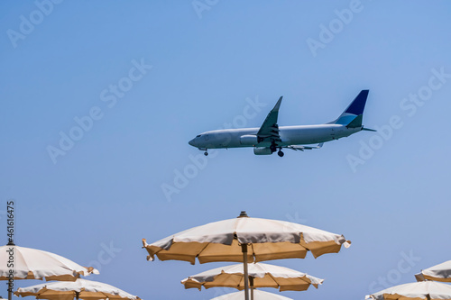 passenger plane flying low above beach with blue sea with resting and swiming tourists and parasol umbrellas on the foreground