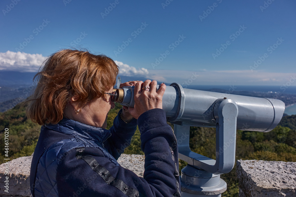 A red-haired woman in glasses and a blue jacket looks through binoculars from a high tower to the sea and the city.