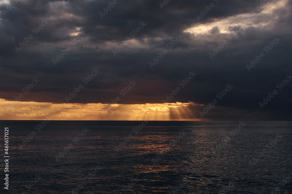 The setting sun illuminates the sea on the horizon with golden light through storm clouds. The sea is storming, it is raining on the right on the horizon.