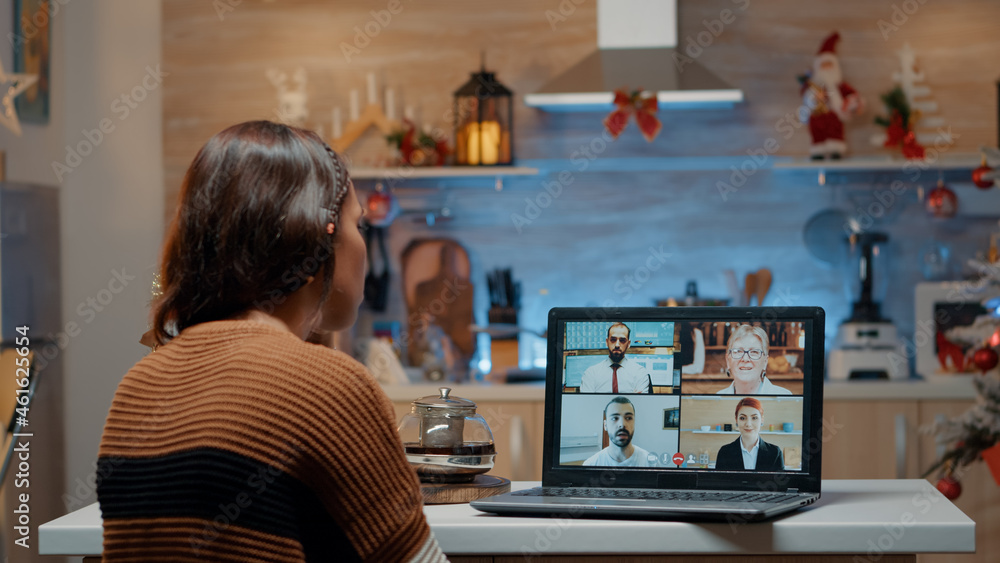 Woman on video call with workmates at home waiting for christmas eve holiday celebration. Young adult sitting in kitchen with ornaments and decorations using laptop to talk to friends Woman on video call with workmates at home waiting for christmas eve holiday celebration. Young adult sitting in kitchen with ornaments and decorations using laptop to talk to friends