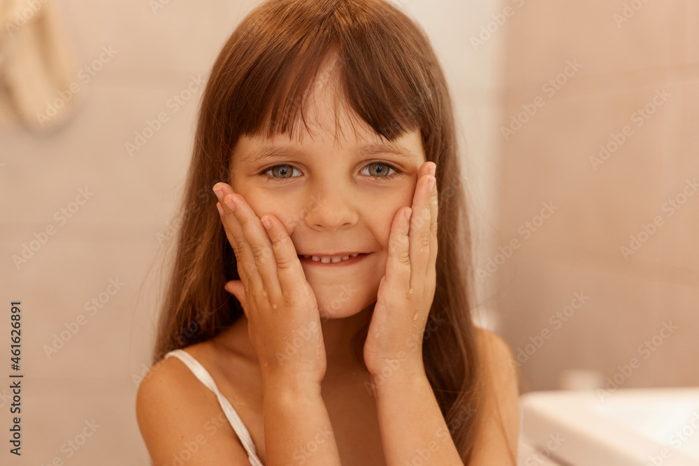 Pretty little girl touching cheeks, looking at camera in with pleasant smile and positive emotions, posing in bathroom after hygienic and beauty procedures. Pretty little girl touching cheeks, looking at camera in with pleasant smile and positive emotions, posing in bathroom after hygienic and beauty procedures.