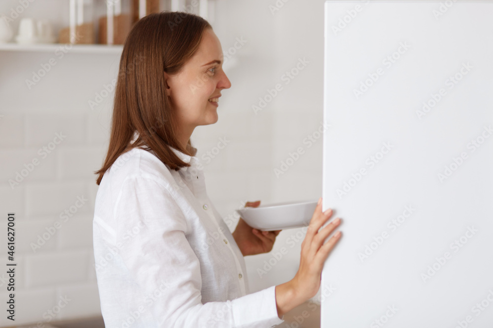 Side view portrait of positive dark haired female wearing white shirt, opening refrigerator, finding food for breakfast or dinner, looking smiling inside fridge. Side view portrait of positive dark haired female wearing white shirt, opening refrigerator, finding food for breakfast or dinner, looking smiling inside fridge.