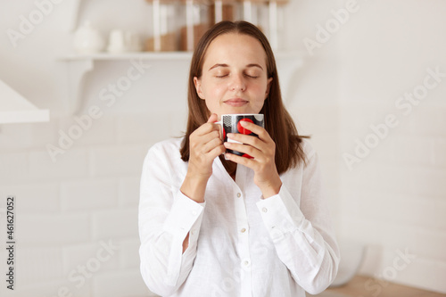 Indoor shot of happy pretty woman enjoying cup of coffee or tea in kitchen in morning, drinking hot beverage, standing with closed eyes with relaxed expression.