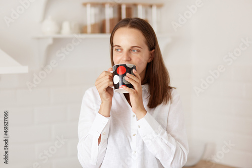 Positive dark haired female wearing white casual style t shirt, drinking coffee or tea in morning, posing with kitchen set of background, looking away with dreamy expression.