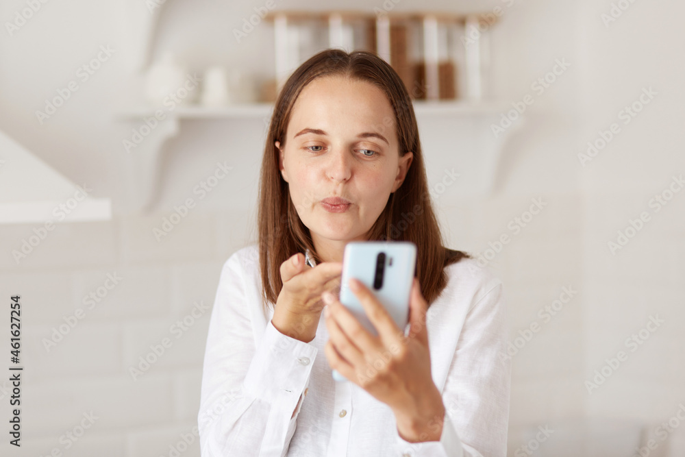 Portrait of young adult happy woman using smartphone for making video calls in the kitchen at home, sending air kissing for followers while broadcasting livestream. Portrait of young adult happy woman using smartphone for making video calls in the kitchen at home, sending air kissing for followers while broadcasting livestream.