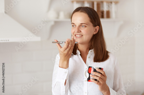 Indoor shot of attractive young adult woman making a voice message with smart phone while relaxing at home and drinking hot tea or coffee  standing in light kitchen.