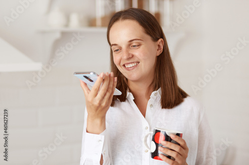 Indoor shot of happy smiling young adult woman standing with smartphone and cup of coffee or tea in hands, wearing white casual style shirt, looking at display of cell phone.