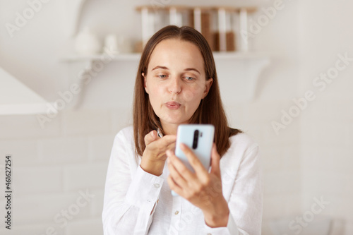 Portrait of young adult happy woman using smartphone for making video calls in the kitchen at home, sending air kissing for followers while broadcasting livestream.