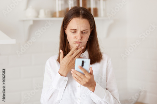 Young adult unhappy woman using smartphone for streaming or having video call, posing in the kitchen at home, wearing white casual style shirt, expressing sad emotions.