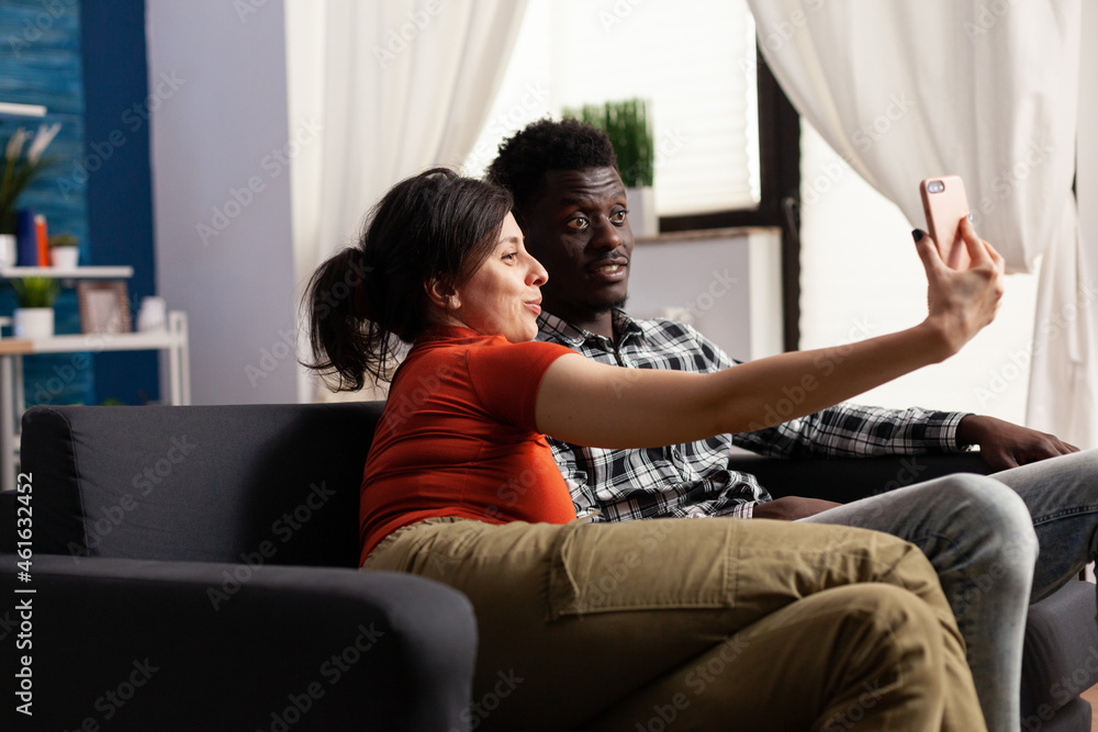 Cheerful interracial couple taking pictures with smartphone at home. Multi ethnic people making memories and selfies with modern device in hand and technology. Mixed race happy partners Cheerful interracial couple taking pictures with smartphone at home. Multi ethnic people making memories and selfies with modern device in hand and technology. Mixed race happy partners