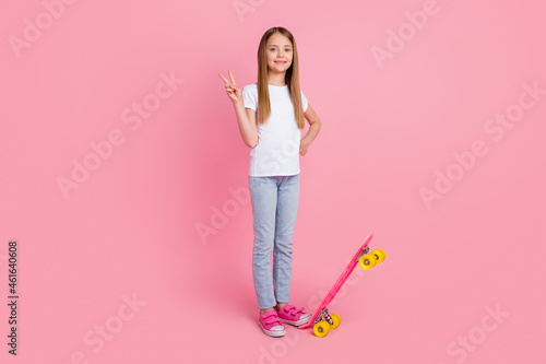 Full size photo of funky blond small girl with skate show v-sign wear white t-shirt jeans isolated on pink color background