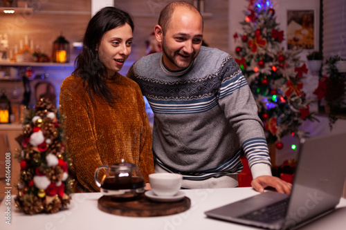 Happy family greeting friends during online videocall meeting on laptop computer celebrating christmas holiday together. Cheerful couple enjoying spending winter season in xmas decorated kitchen