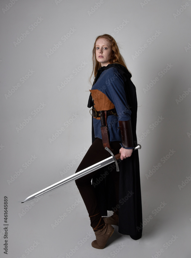 Full length, portrait of red haired woman wearing medieval viking inspired costume and flowing cloak, Holding a long sword weapon posing against studio background. Full length, portrait of red haired woman wearing medieval viking inspired costume and flowing cloak, Holding a long sword weapon posing against studio background.