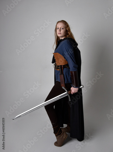 Full length, portrait of red haired woman wearing medieval viking inspired costume and flowing cloak,  Holding a long sword weapon  posing against studio background. photo