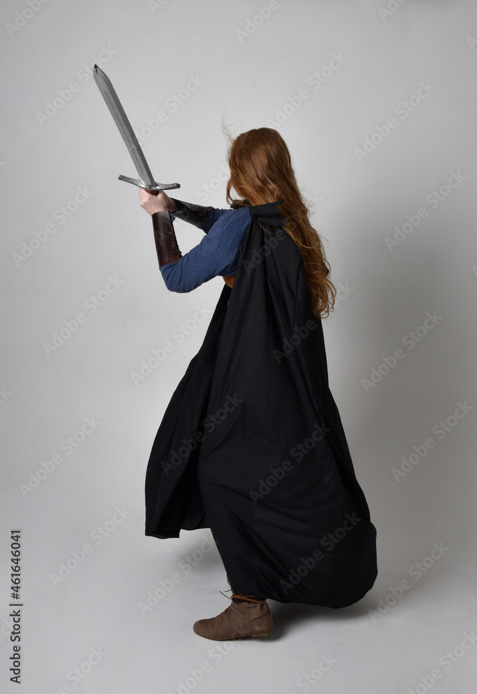 Full length, portrait of red haired woman wearing medieval viking inspired costume and flowing cloak, Holding a long sword weapon posing against studio background. Full length, portrait of red haired woman wearing medieval viking inspired costume and flowing cloak, Holding a long sword weapon posing against studio background.