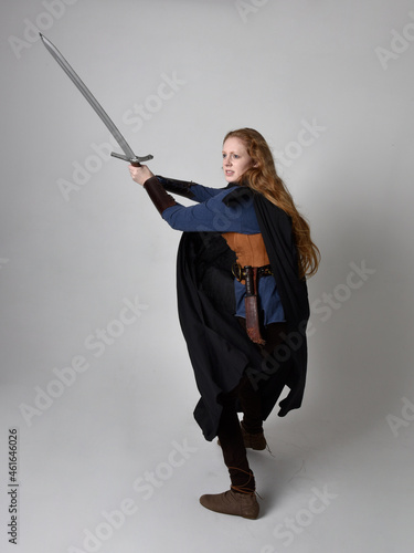 Full length, portrait of red haired woman wearing medieval viking inspired costume and flowing cloak, Holding a long sword weapon posing against studio background.