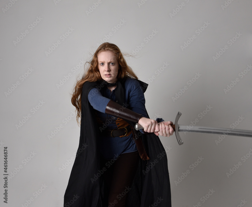 Close up portrait of red haired woman wearing medieval viking inspired costume and flowing cloak, Holding a long sword weapon, action pose against studio background. Close up portrait of red haired woman wearing medieval viking inspired costume and flowing cloak, Holding a long sword weapon, action pose against studio background.