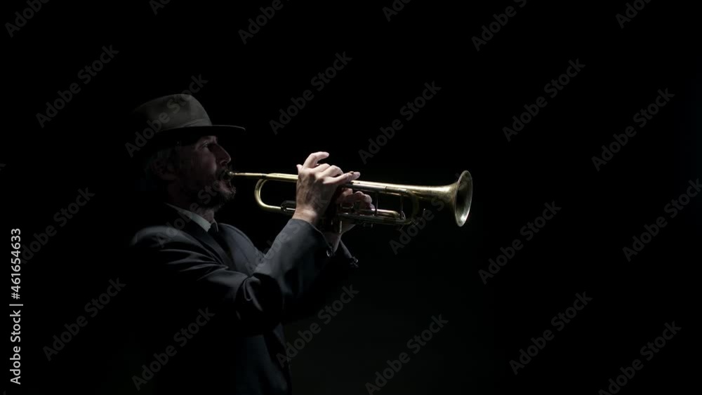 Man in jacket and hat picks up trumpet and plays music on it at black background, side view. Man plays blues on trumpet showing master class