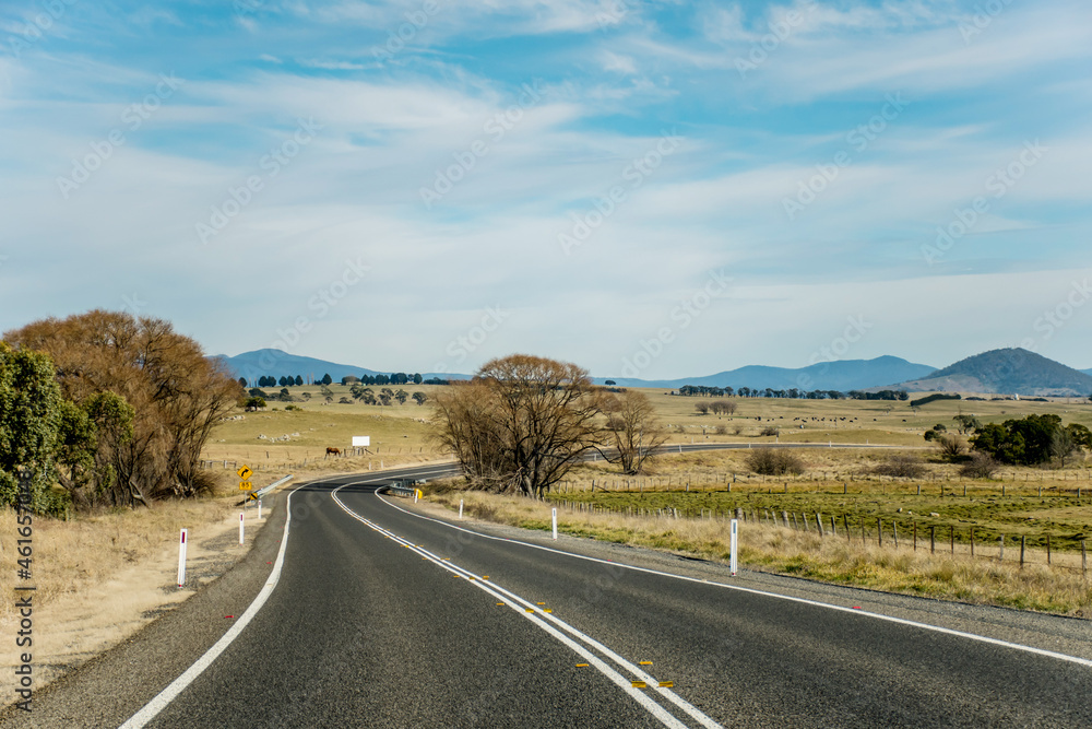 Open empty road surrounded by farms and fields in Australia. Mountains on the horizon. Road trip travel