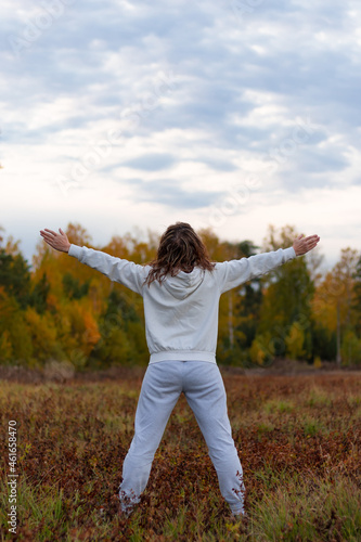 A woman in a white sweater with a hood stands in a field against the background of a forest and a blue sky with her arms outstretched wide on a warm autumn day. Selective focus. Close-up. Portrait