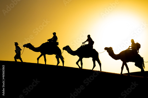 A caravan of camels with tourists moves through the dunes of the Sahara desert at sunset