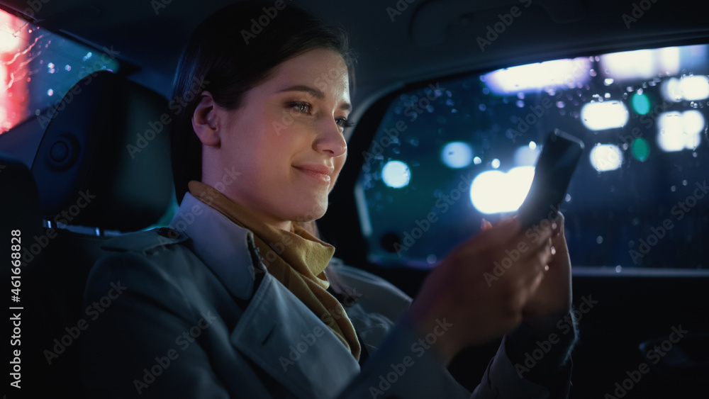 Stylish Female is Commuting Home in a Backseat of a Taxi at Night. Beautiful Woman Passenger Using Smartphone and Looking Out of Window while in a Car in Urban City Street with Working Neon Signs. - obrazy, fototapety, plakaty 