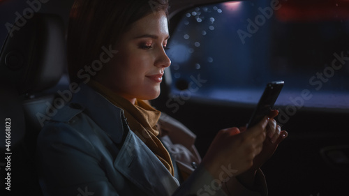 Stylish Female is Commuting Home in a Backseat of a Taxi at Night. Beautiful Woman Passenger Using Smartphone and Looking Out of Window while in a Car in Urban City Street with Working Neon Signs.