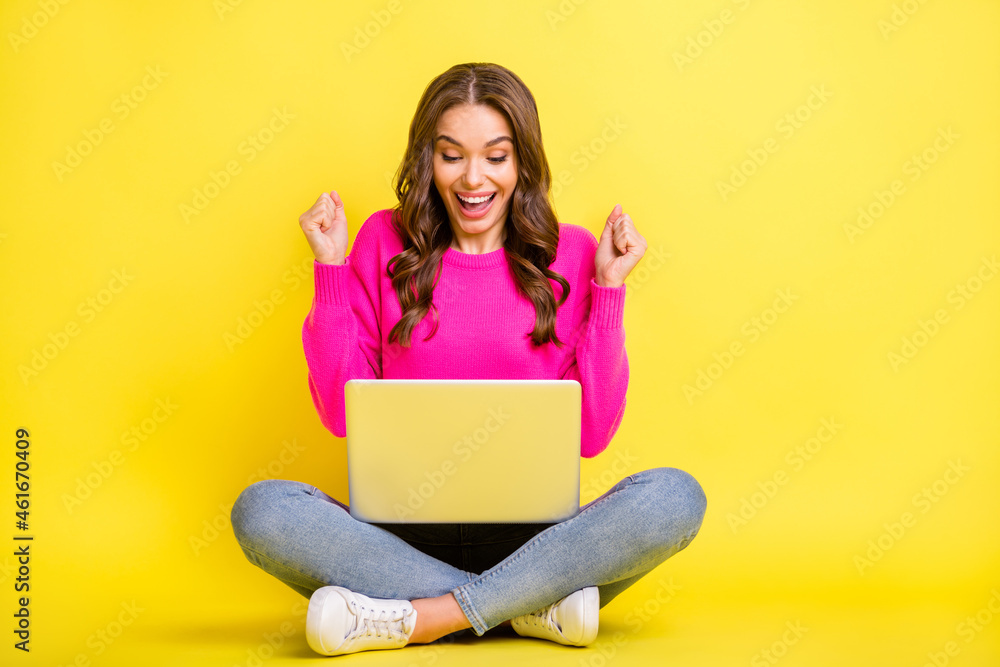 Full size photo of young happy excited crazy smiling girl sit floor look laptop hold fists in victory isolated on yellow color background Full size photo of young happy excited crazy smiling girl sit floor look laptop hold fists in victory isolated on yellow color background