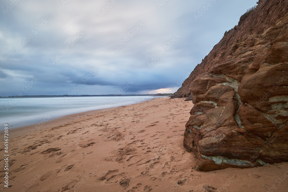 Scenic view of a sandy beach landscape with volcanic rocks in the foreground and ocean in the background, cloudy blue sky above the horizon at Phillip Island, Australia.
