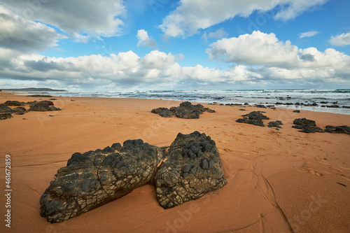 Scenic view of a sandy beach landscape with volcanic rocks in the foreground and ocean in the background, cloudy blue sky above the horizon at Phillip Island, Australia. 