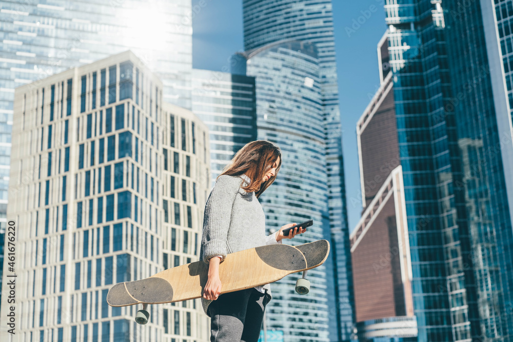 Portrait of casually dressed young businesswoman with skateboard using phone near office.