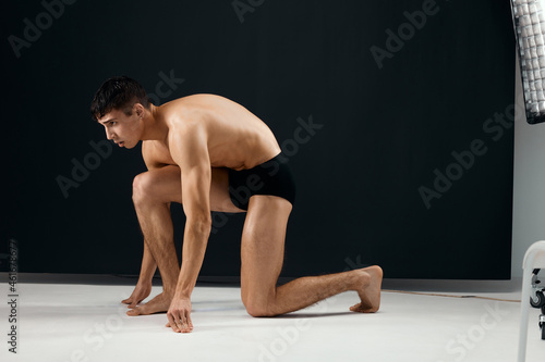 male bodybuilder in black shorts stands on his knee against a dark background
