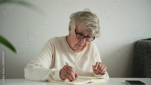Elderly woman with stylish grey short haircut in white woolen jacket writes down autobiography for future generation in paper notebook photo