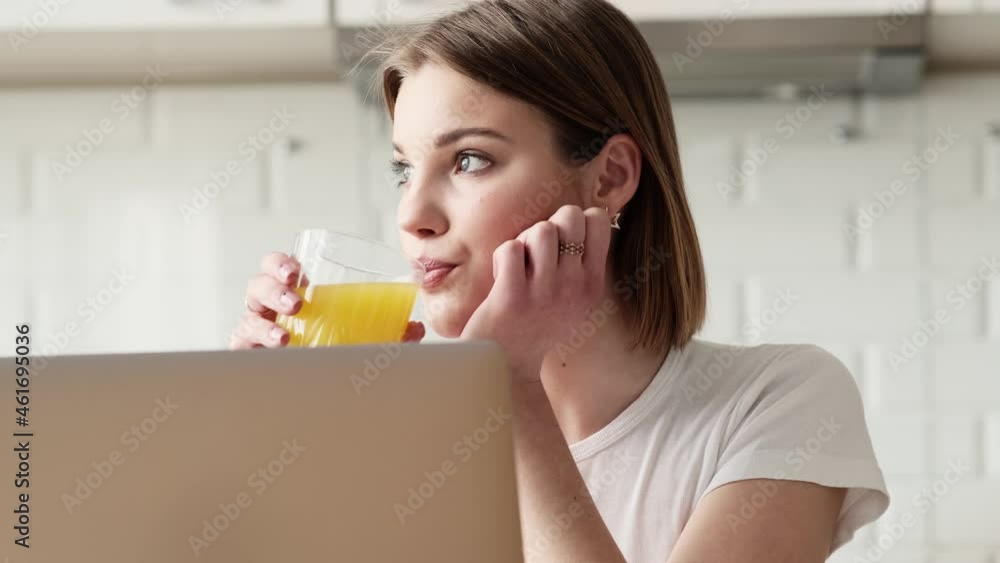 A pleased woman is using her laptop computer while drinking orange juice sitting inside the apartments A pleased woman is using her laptop computer while drinking orange juice sitting inside the apartments