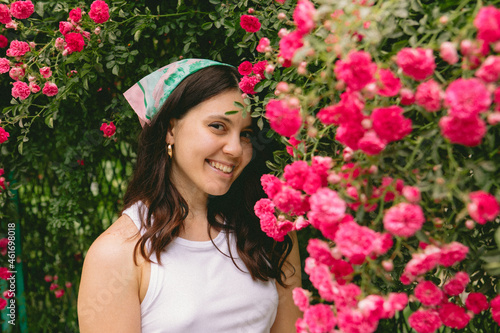 young pretty smiling woman near blooming red roses bush