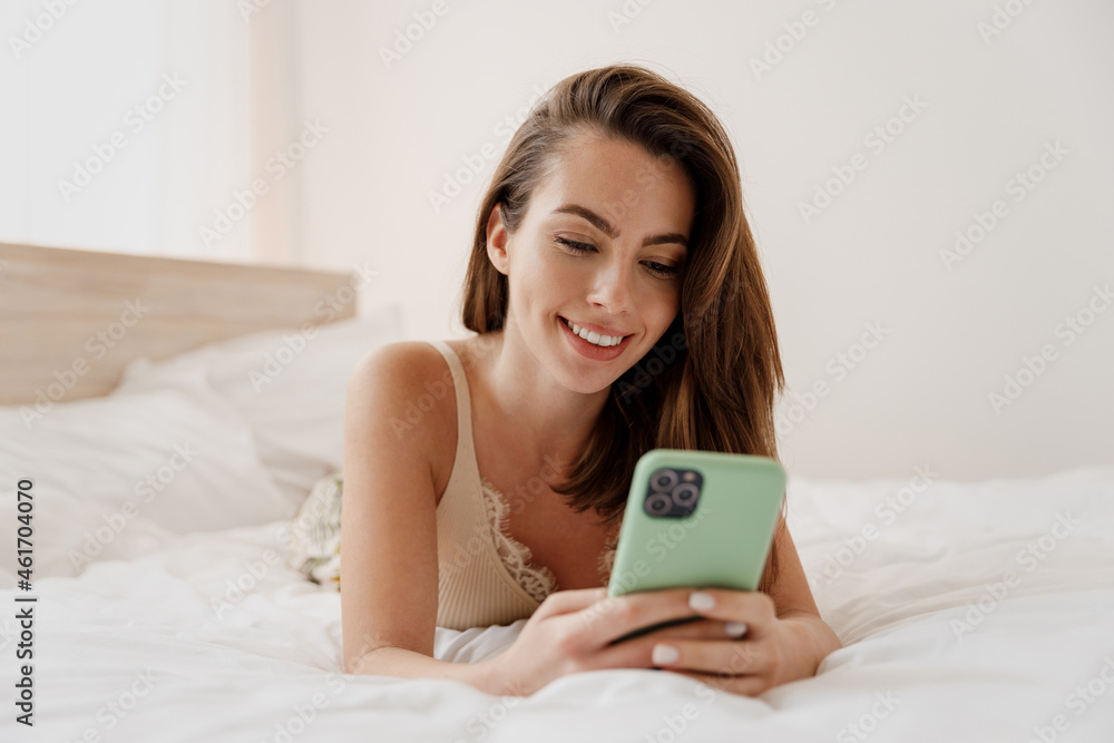 Beautiful happy young woman in casual casual clothes lying on her stomach hold in hand use mobile cell phone rest relax spend time in bedroom lounge home in own room house be lost in reverie good day. Beautiful happy young woman in casual casual clothes lying on her stomach hold in hand use mobile cell phone rest relax spend time in bedroom lounge home in own room house be lost in reverie good day.