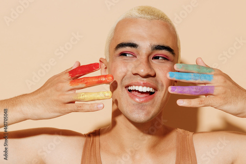 Young smiling curious minded happy fun blond latin gay man with make up fingers painted in rainbow flag color wear beige tank shirt look aside isolated on plain light ocher background studio portrait