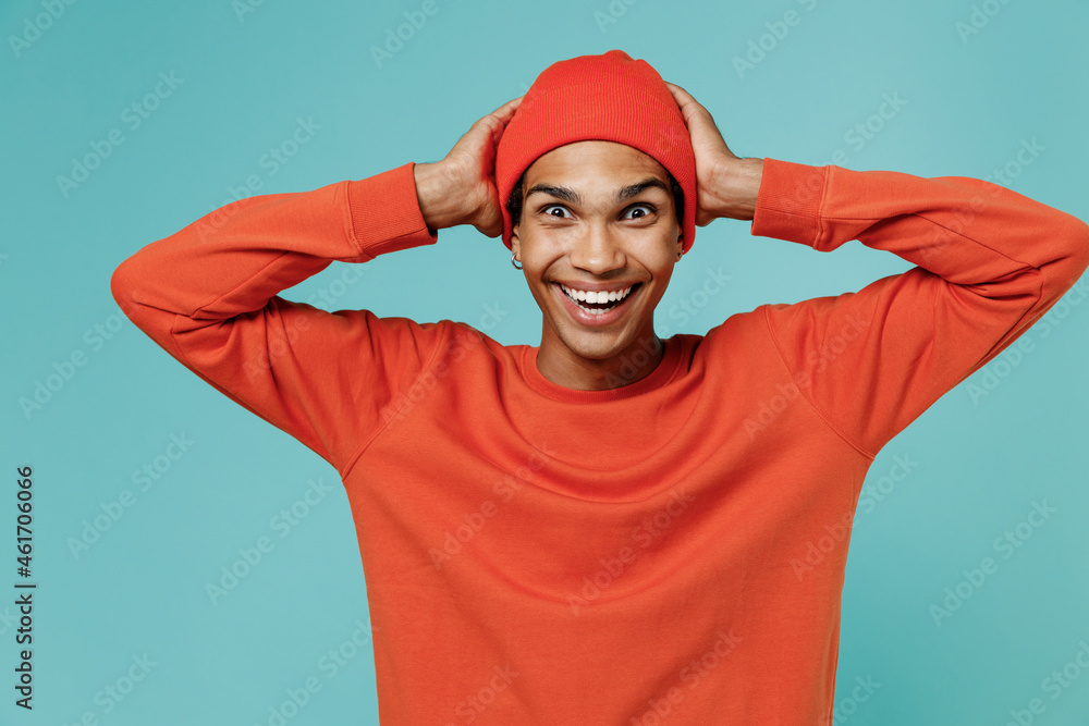 Fototapeta premium Young surprised shocked amazed happy cool smiling african american man 20s wearing red shirt hat hold head isolated on plain pastel light blue background studio portrait. People lifestyle concept