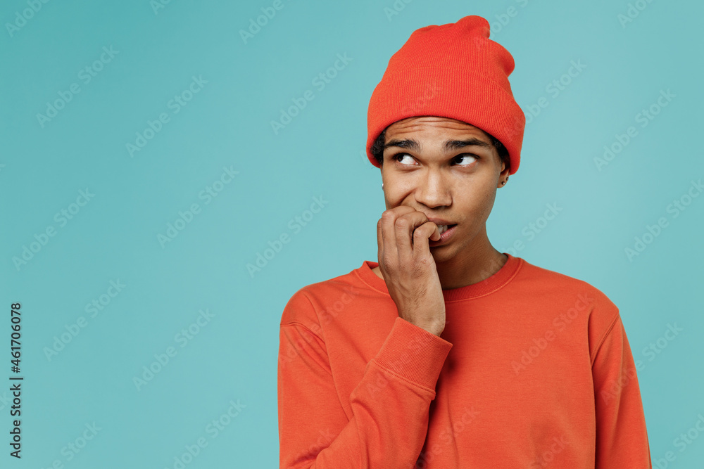 Young sad puzzled confused embarrassed african american man 20s in orange shirt hat look aside biting fingers isolated on plain pastel light blue background studio portrait. People lifestyle concept.