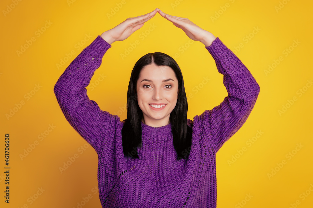 Young smiling happy girl holding folded hands above head like roof home house relocation yellow background studio portrait