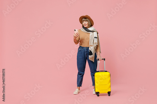 Full size traveler tourist vivid mature elderly woman 55 years old wear casual clothes hat scarf hold suitcase bag use mobile cell phone isolated on plain pastel light pink background studio portrait