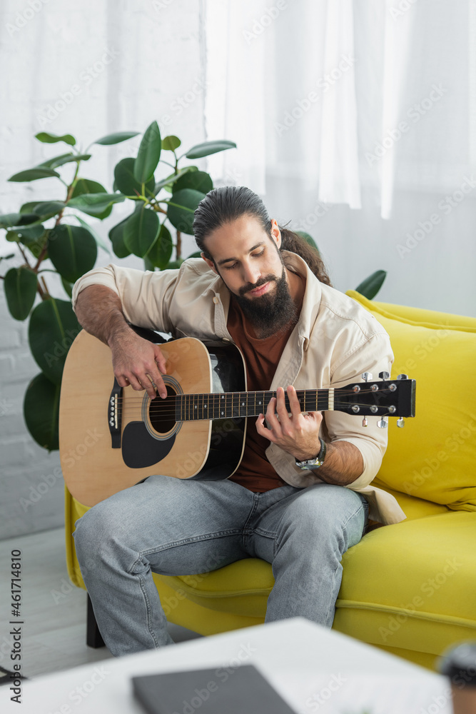 bearded latin man playing acoustic guitar while sitting on yellow couch at home