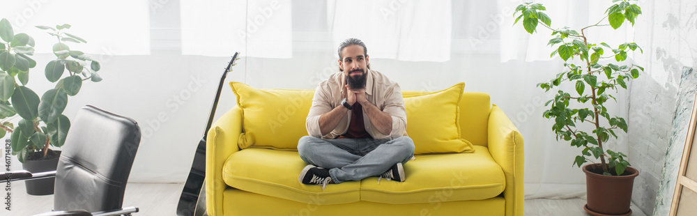 bearded hispanic man meditating on yellow couch in living room, banner