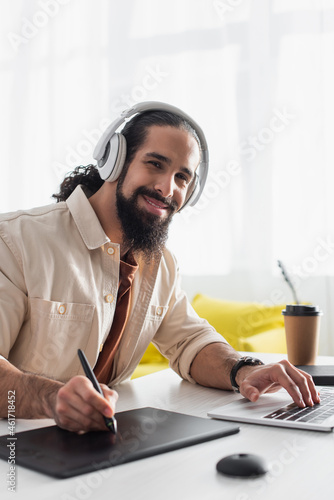 smiling hispanic designer in headphones working on laptop and drawing tablet at home