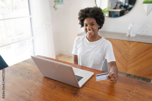 Smiling young African female entrepreneur working online with a laptop while sitting at her kitchen table at home