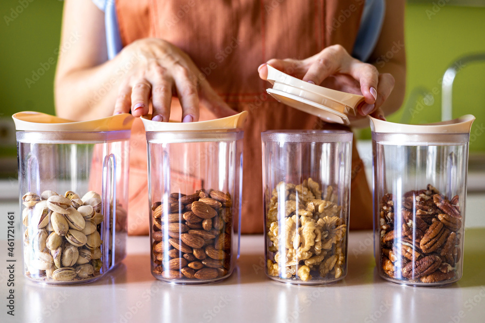 Closeup woman hands in apron placing different nuts into glass jar storage container at kitchen