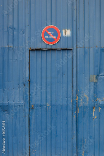 Postão metálico antigo de armazém em zona industrial, com aspeto abandonado e danificado, pintado na cor azul e com o sinal de trânsito de proibição de estacionar photo