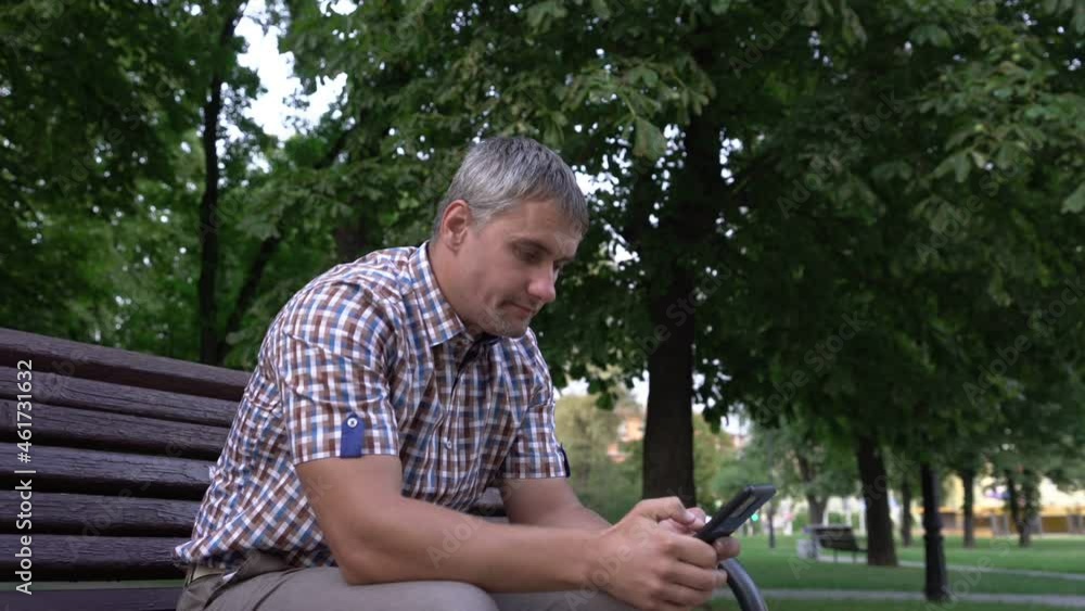 Mature man with grey hair dressed in checkered shirt browses social networks on phone sitting on wooden bench in large city park Mature man with grey hair dressed in checkered shirt browses social networks on phone sitting on wooden bench in large city park